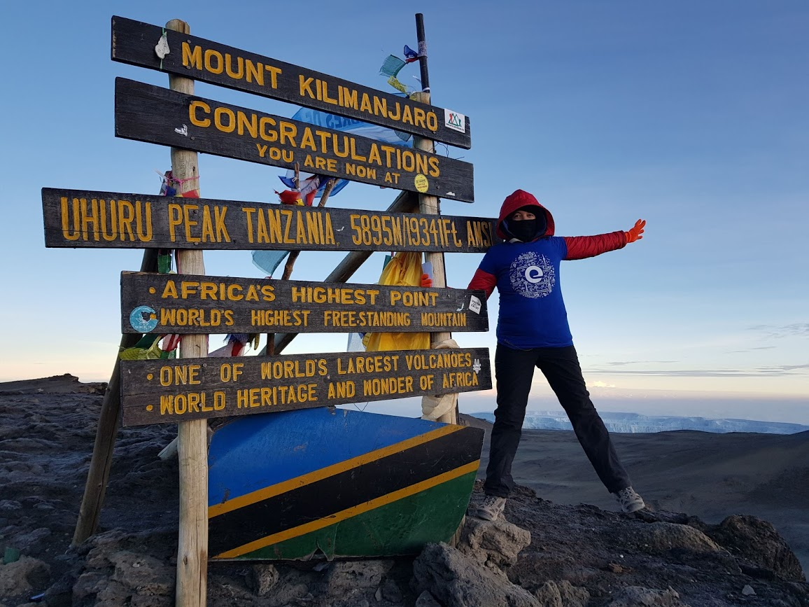person wearing blue shirt and black pants posing next to the Mount Kilimanjaro peak sign