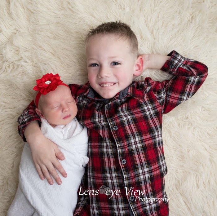 Young boy smiling at camera wearing plaid red shirt hugging small sleeping baby wearing red headband