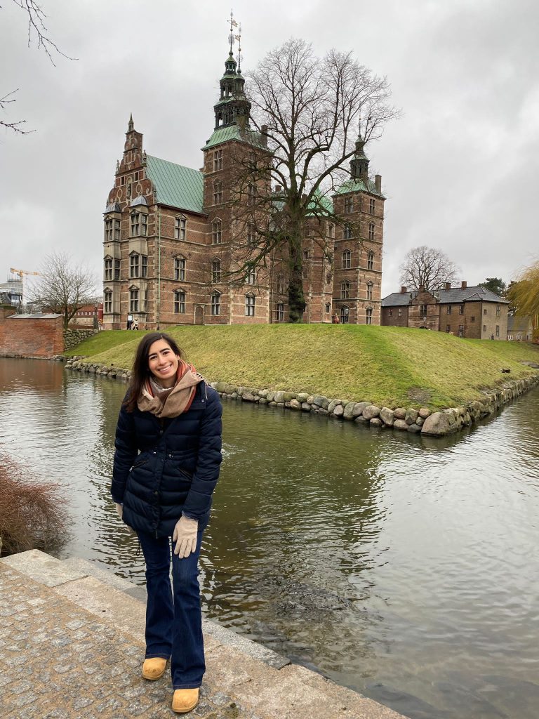 Sonia, wearing a dark blue jacket,  standing by a river, with a historical style building behind. 
