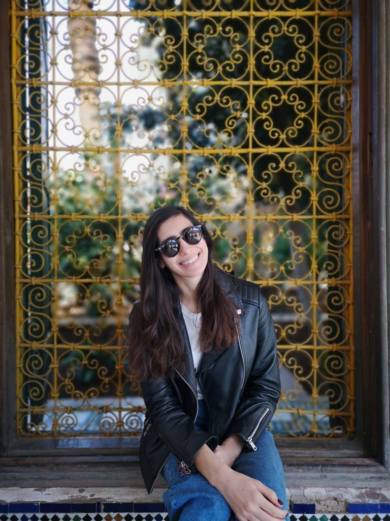 Sonia sitting in front of a patterned window, wearing sunglasses and a black jacket.