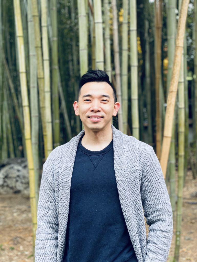 Marc outdoors, wearing a grey cardigan and dark blue top, with bamboo trees in the background.